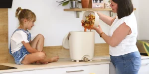 Un'immagine che mostra una signora che versa i suoi rifiuti alimentari dal Kitchen caddy nel bidone bokashi, che si trova sul lato della cucina. La figlia è seduta accanto al bidone bokashi sul bancone della cucina.